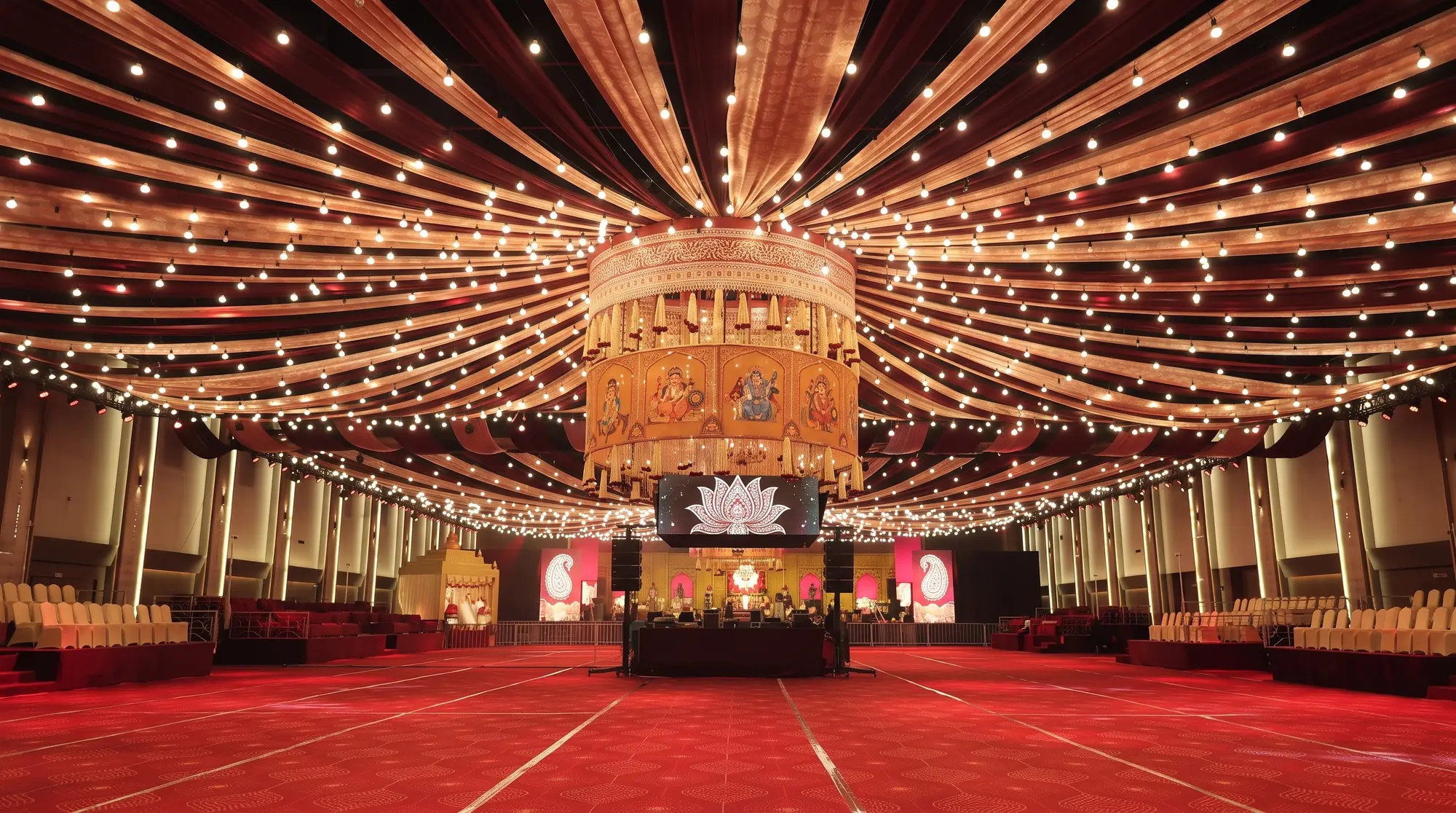 Vast event hall with a fully draped ceiling under thousands of bulbs, a large circular chandelier with painted panels, an LED lotus screen and a decorated stage