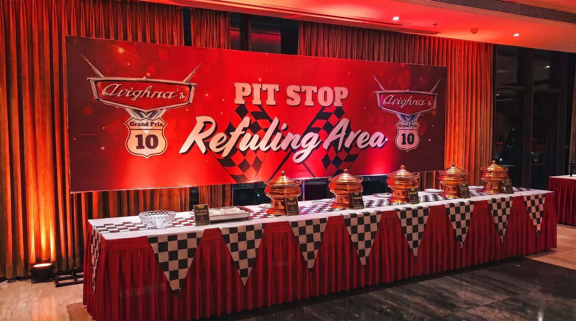 Catering counter dressed as a pit stop refuelling area, with a printed backdrop, brass chafing dishes and chequered bunting at a 10th birthday in Lake Town, Kolkata